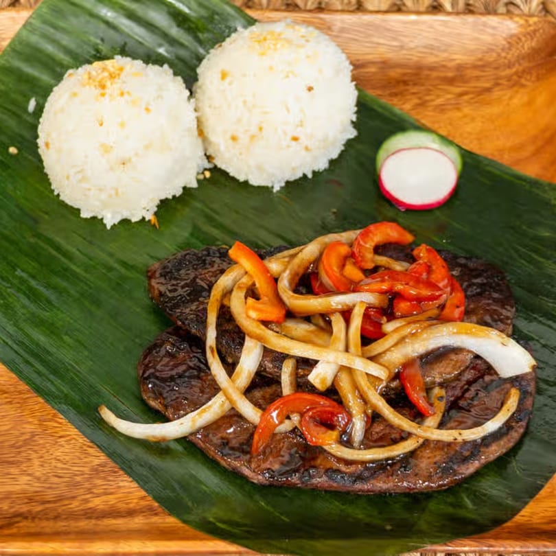 Filipino bistek with onions and red bell peppers, two scoops of rice, and a radish slice on a banana leaf.