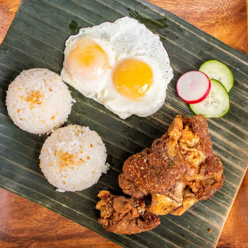 Filipino meal with crispy pata, two fried eggs, garlic rice, and sliced radishes and cucumbers on a banana leaf.