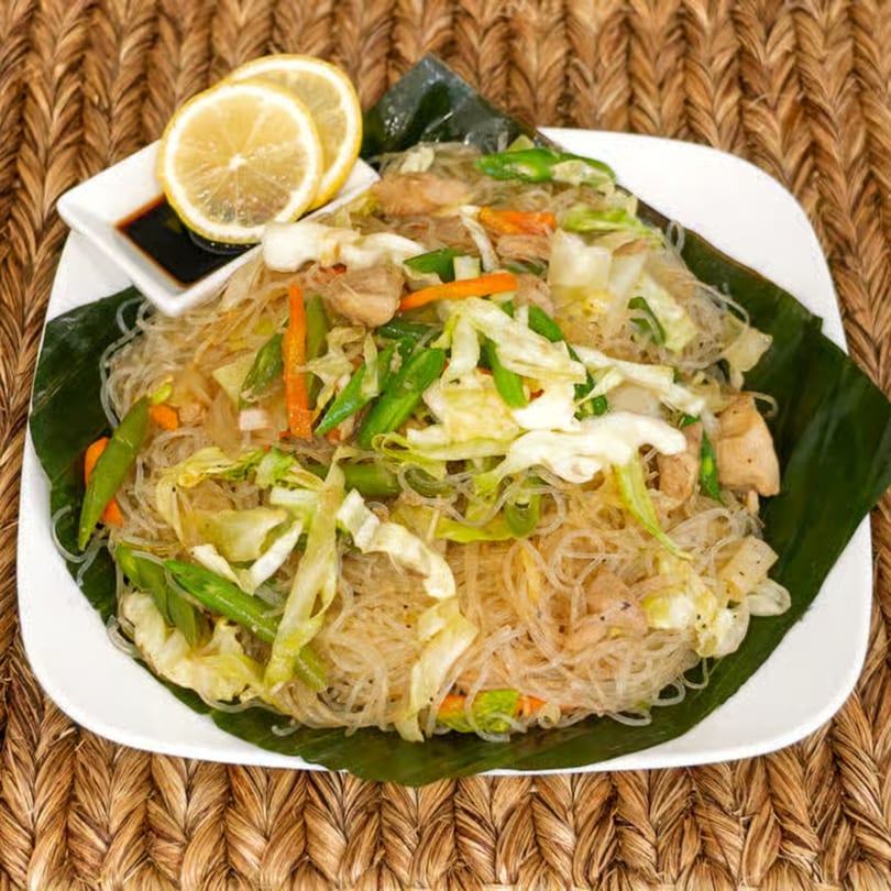 Filipino Pancit Bihon with chicken, vegetables, and lemon slices on a banana leaf-lined plate.